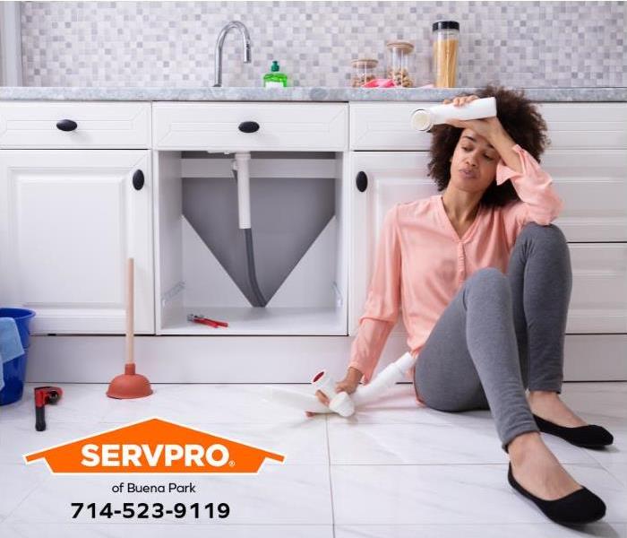 A person sits on the kitchen floor holding a broken plumbing pipe.